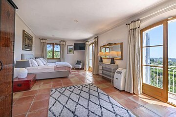 Sunny bedroom with terracotta floor, large bed, and balcony overlooking green landscape. Includes a decorative rug, mirror, and TV.