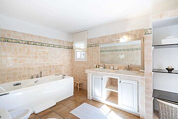 Spacious bathroom with light peach tiled walls, a white bathtub, double vanity, large mirror, and terracotta floor. Features a decorative border and shelves.