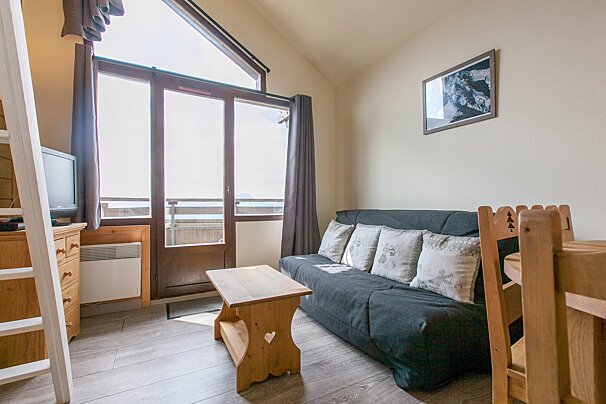 Bright studio apartment with a dark sofa, wooden furniture, and large windows leading to a balcony, filled with natural light.