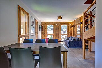 Modern chalet interior featuring a dining table with dark chairs, wooden walls, and a cozy living area with a blue sofa.