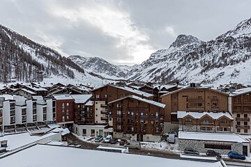 A snowy mountain village with chalet-style buildings, surrounded by vast, snow-covered peaks and sparse trees under a cloudy sky.