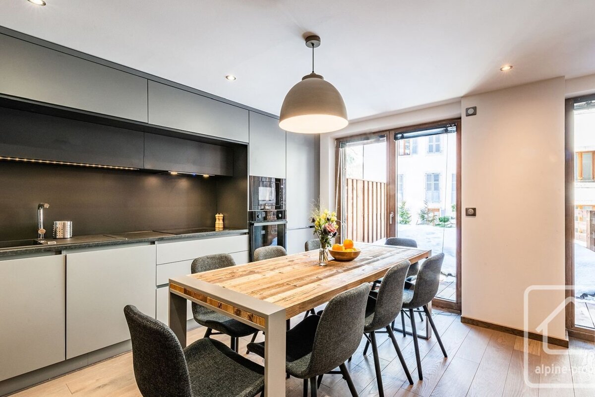 A modern kitchen and dining area with sleek grey cabinets, a rustic wooden table, grey chairs, and large glass doors providing natural light.