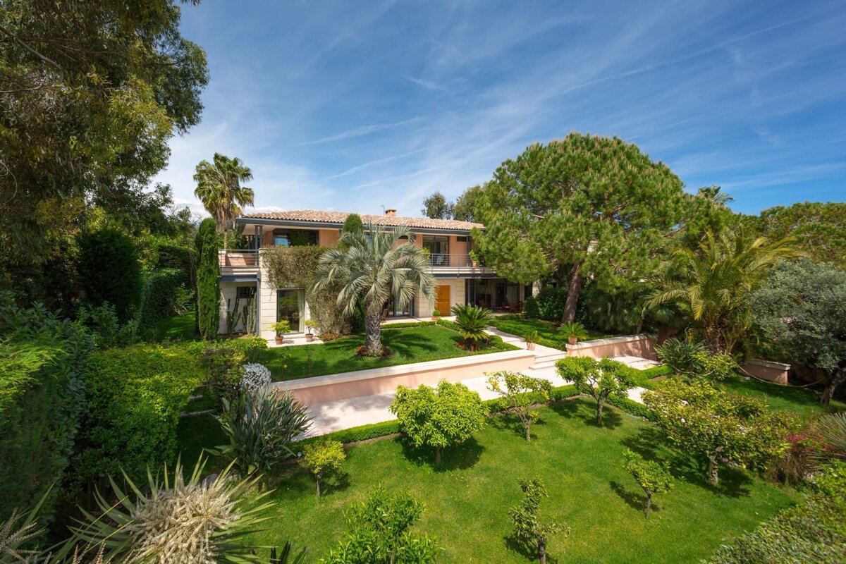 Large two-story villa with a lush green lawn, palm trees, and landscaped gardens under a bright blue sky.