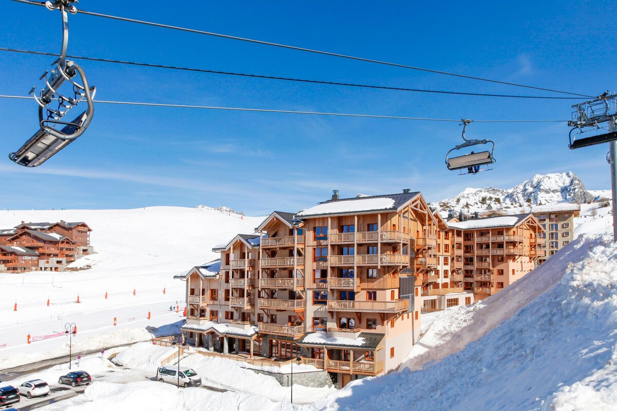 A sunny ski resort with modern wooden chalets, snowy slopes, and chairlifts overhead against a clear blue sky and distant mountains.