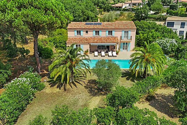 Aerial view of a peach Mediterranean villa with blue shutters, a tiled roof, and a swimming pool, surrounded by palm trees and lush green foliage.