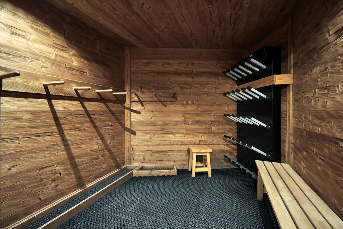 A rustic, wood-paneled ski boot drying room with wall-mounted boot dryers, a bench, a stool, and a textured floor.