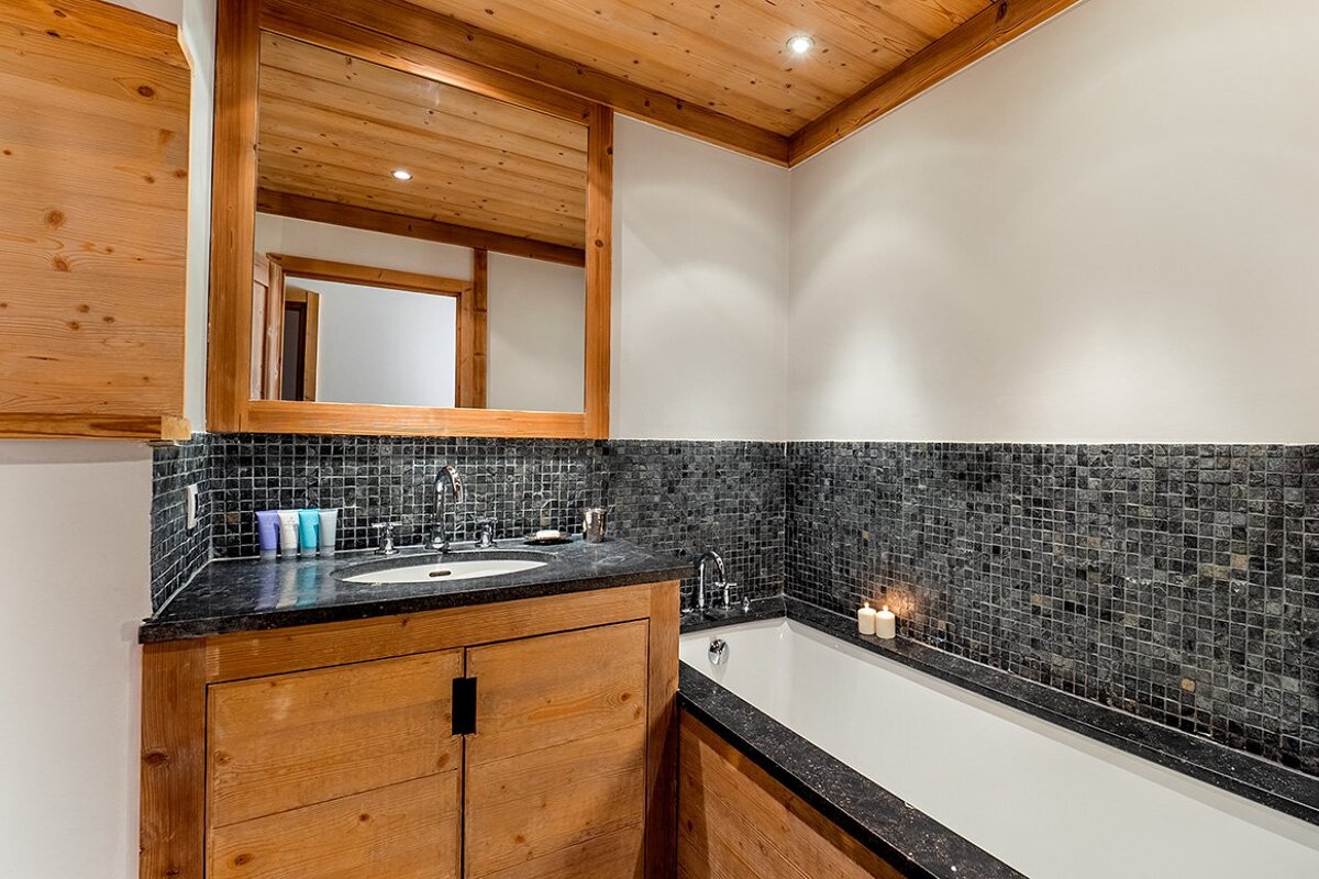 Cozy bathroom featuring a wooden vanity and ceiling, black mosaic tile walls, a large mirror, and a built-in bathtub.