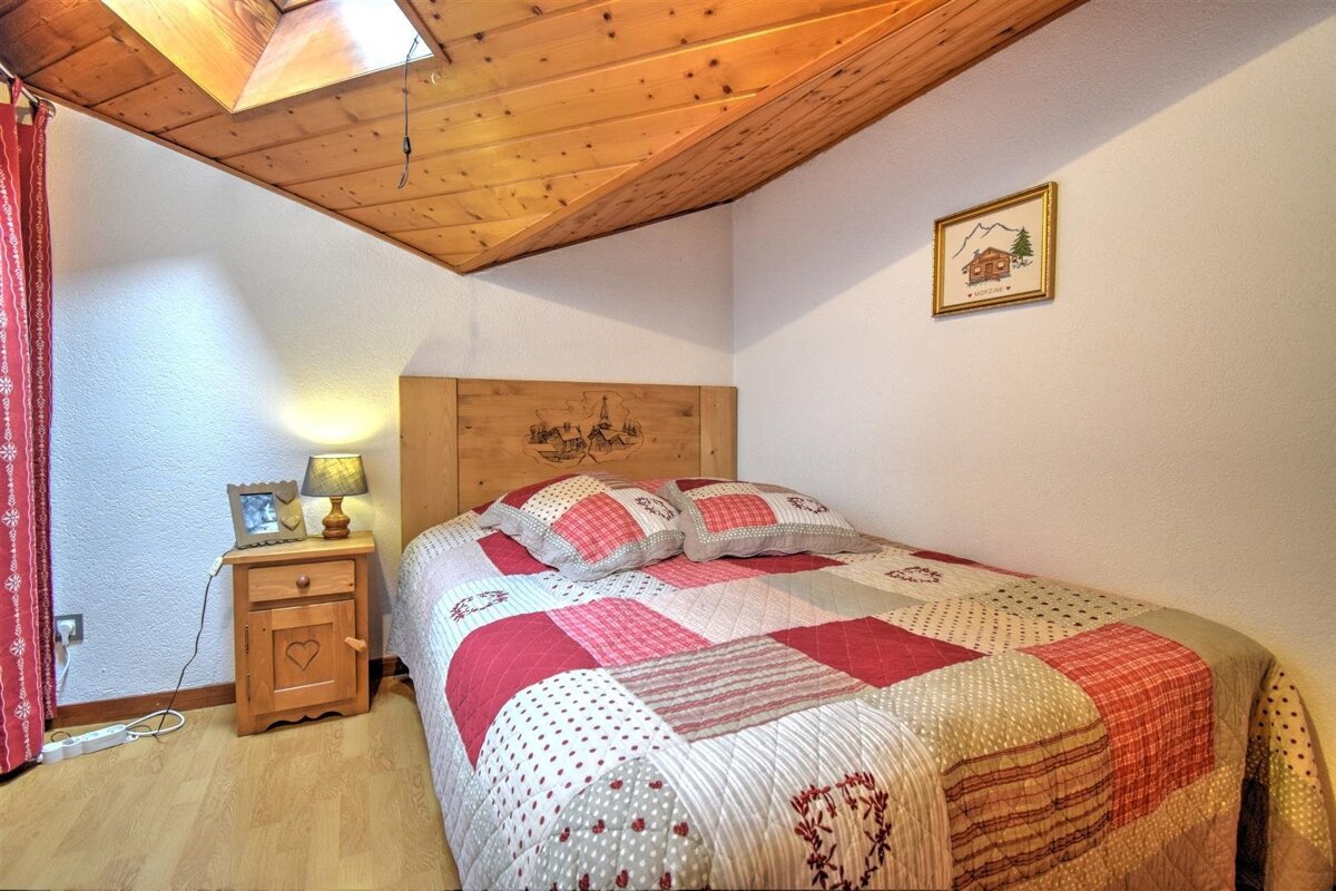 Cozy bedroom with a red patchwork quilt, rustic wooden headboard, sloped wood ceiling with a skylight, and a bedside lamp.