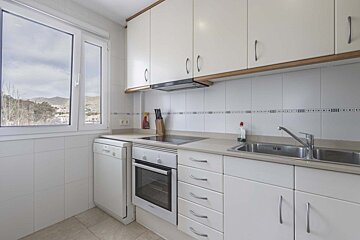 Bright, clean white kitchen with modern appliances (dishwasher, oven, sink), upper/lower cabinets, and a large window offering a scenic mountain view.