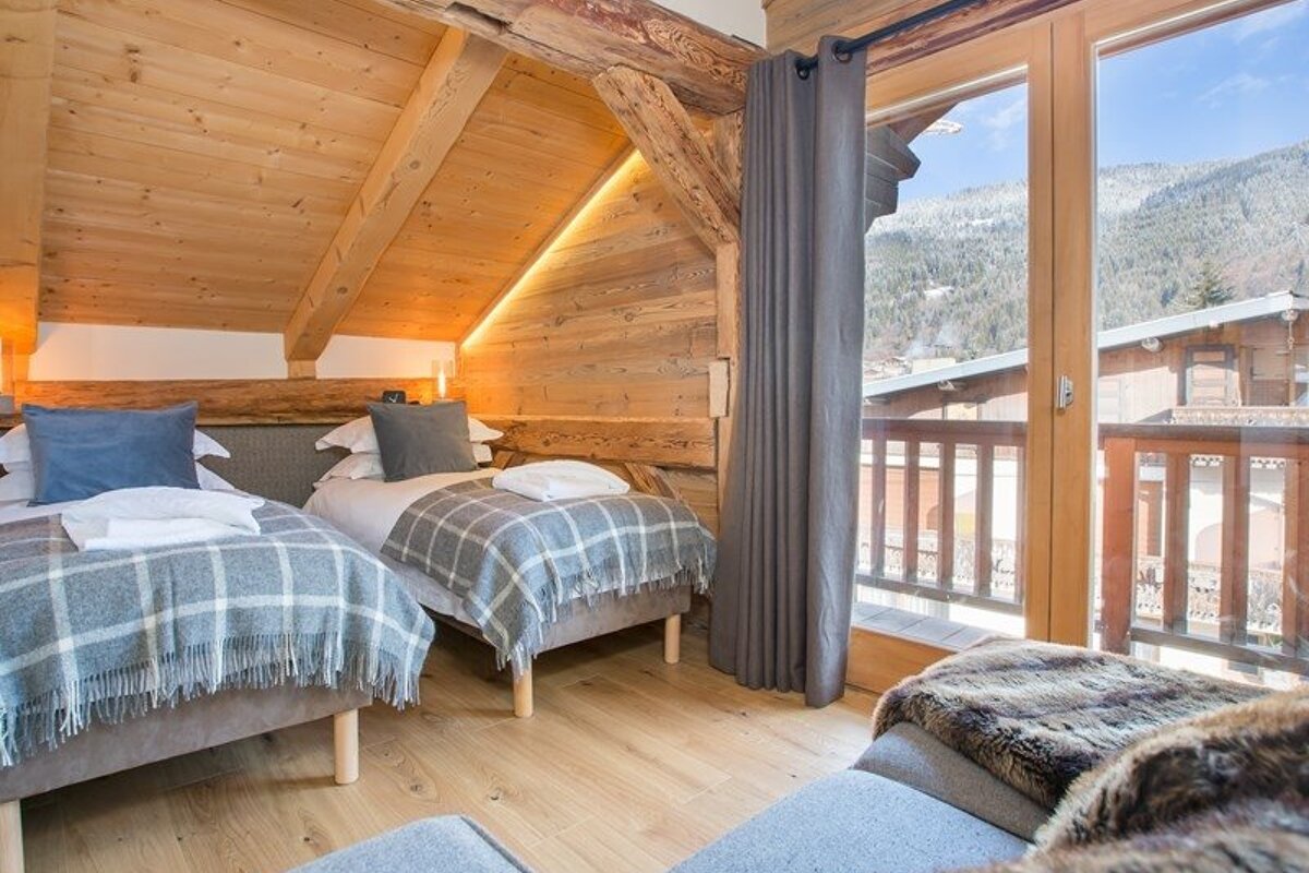 Cozy rustic chalet bedroom featuring two twin beds, wooden interior, and large glass doors with a scenic mountain view.