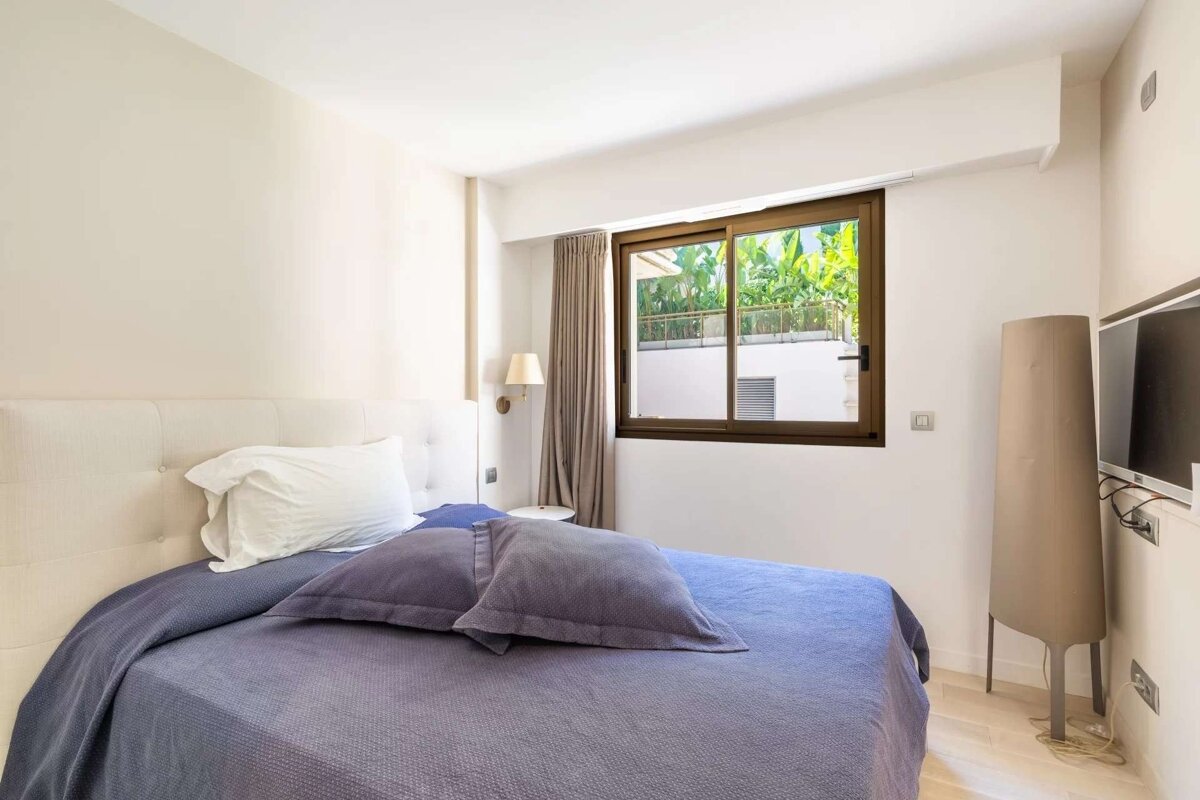 A modern bedroom featuring a bed with purple bedding and a white headboard. A large window offers a view of lush greenery. A TV is mounted on the wall.