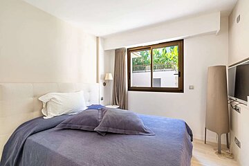 Modern bedroom with a bed, white headboard, purple linens, a window showcasing outdoor greenery, a wall-mounted TV, and a floor lamp.