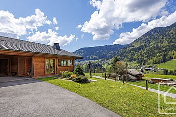 A charming wooden house with a paved driveway and green yard, nestled in a vibrant mountain valley with a distant village under a bright, cloudy sky.