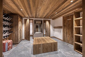 Chalet-style space with wood-paneled ceiling & walls. Features a wine rack, mirror, central wooden island, & storage on a grey stone floor.