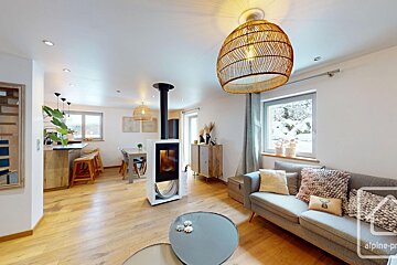 Bright, modern open-plan living space with a cozy fireplace, rattan chandeliers, wooden floors, and a kitchen/dining area. Snow is visible outside.