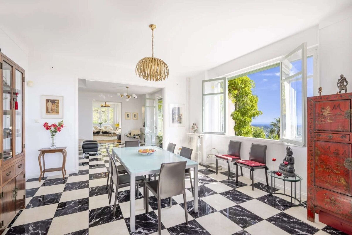 A bright, eclectic dining room with black & white checkered floor, modern table, and open window offering lush green views to the sea.