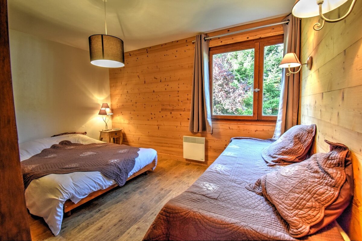 A cozy bedroom with two beds, wood-paneled walls, and a window looking out onto greenery. Warm lighting illuminates the space.