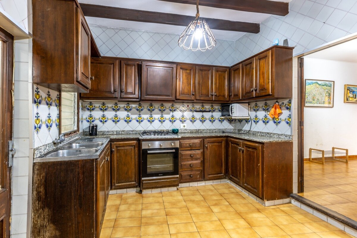 A kitchen with dark wooden cabinets, a vibrant yellow and blue tiled backsplash, and light brown floor tiles. It features a sink, oven, and microwave, opening to another room.