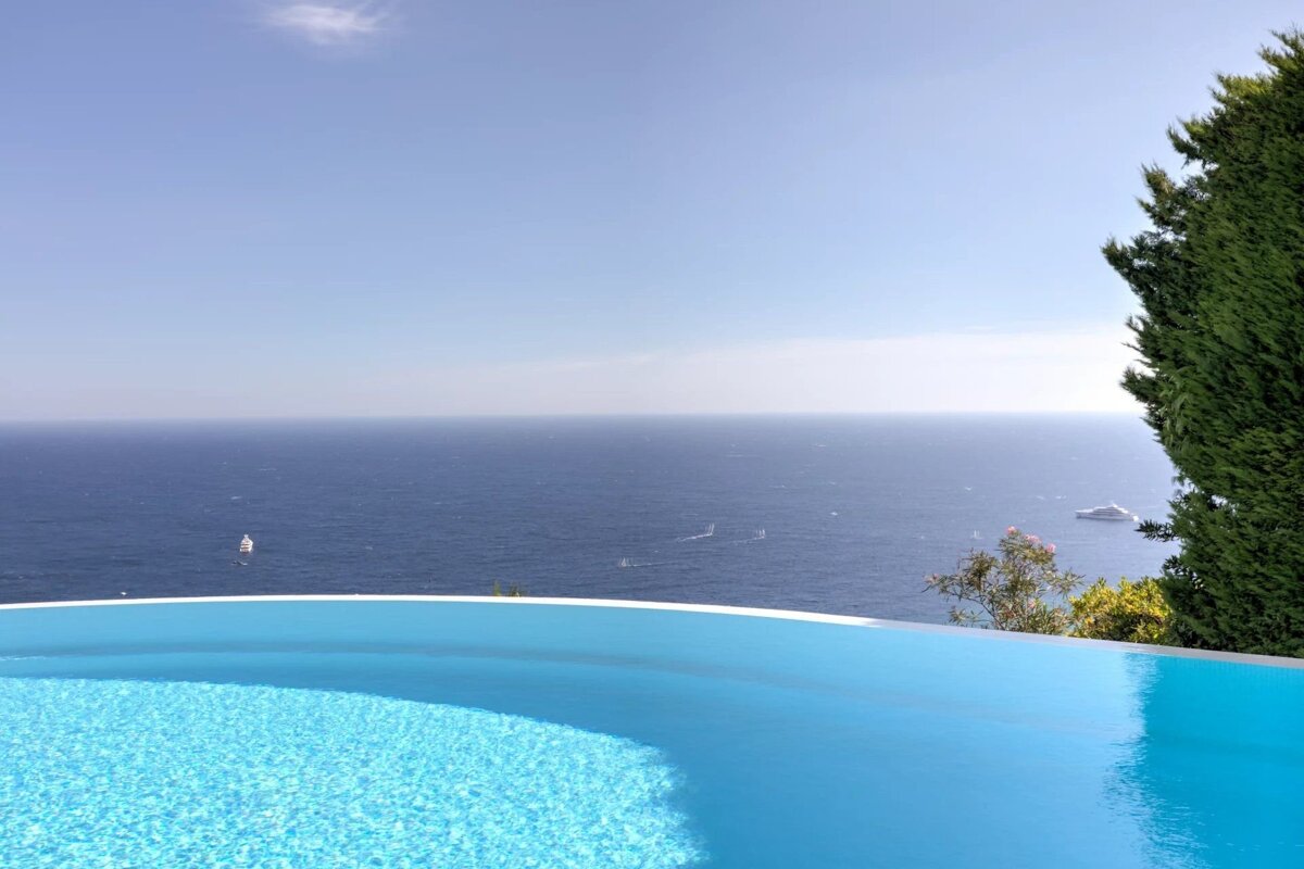 An infinity pool overlooks a deep blue ocean under a clear sky, with boats in the distance.