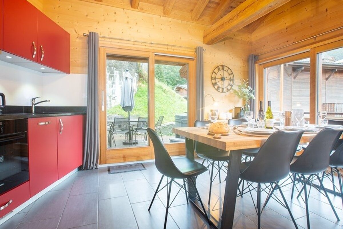 Modern chalet kitchen and dining area featuring vibrant red cabinets, a wooden table with black chairs, and large glass doors.