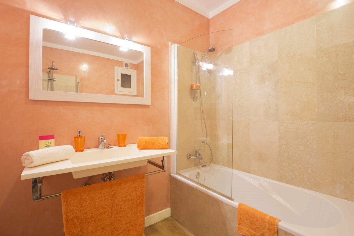 A peach-walled bathroom features a white sink with orange accents, a large mirror, and a bathtub with a glass shower screen.