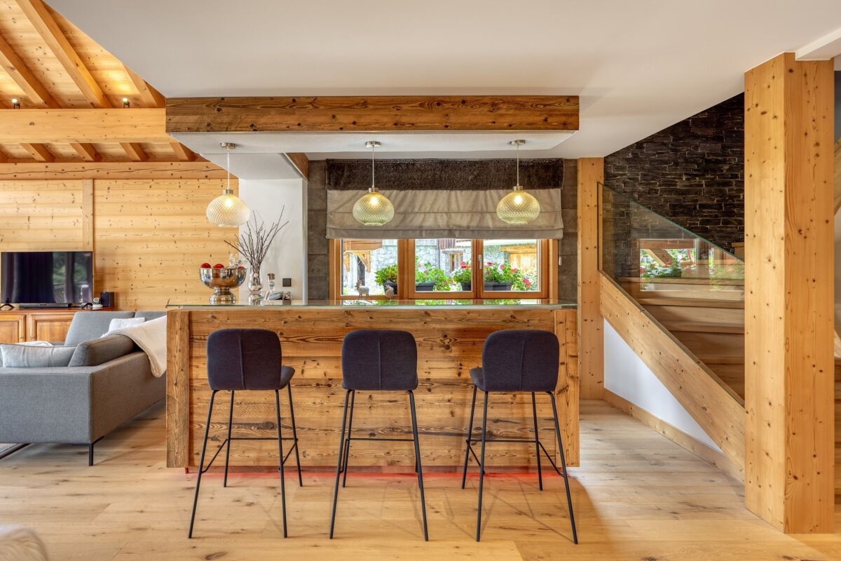 Modern wooden chalet interior featuring a bar with three dark stools, pendant lights, a cozy living area, and a staircase.