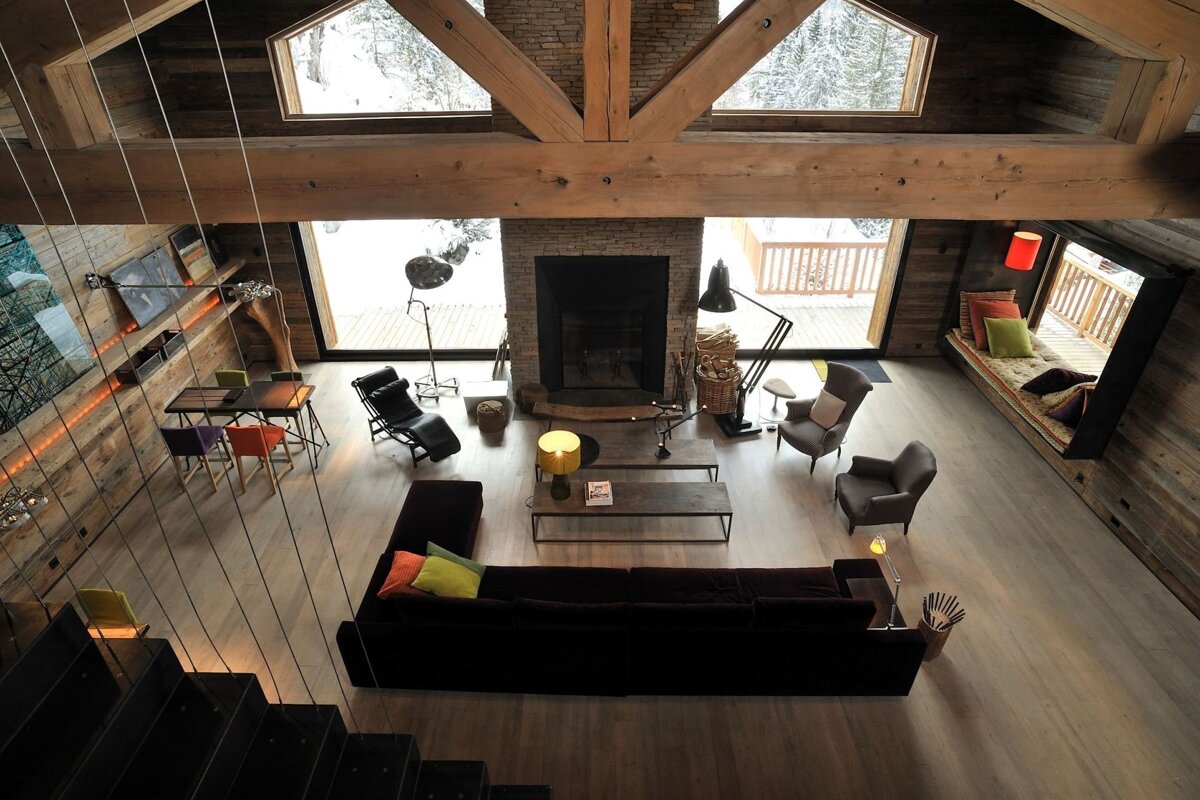 Overhead view of a luxurious, modern rustic living space with high wood-beamed ceilings, fireplace, and large windows revealing a winter landscape.