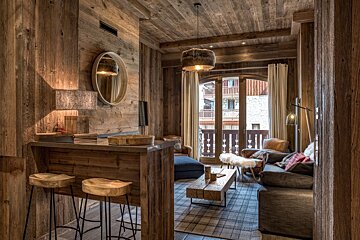 Inviting rustic ski chalet interior with a wooden bar, stools, plush seating in the living room, and a balcony view of alpine buildings.