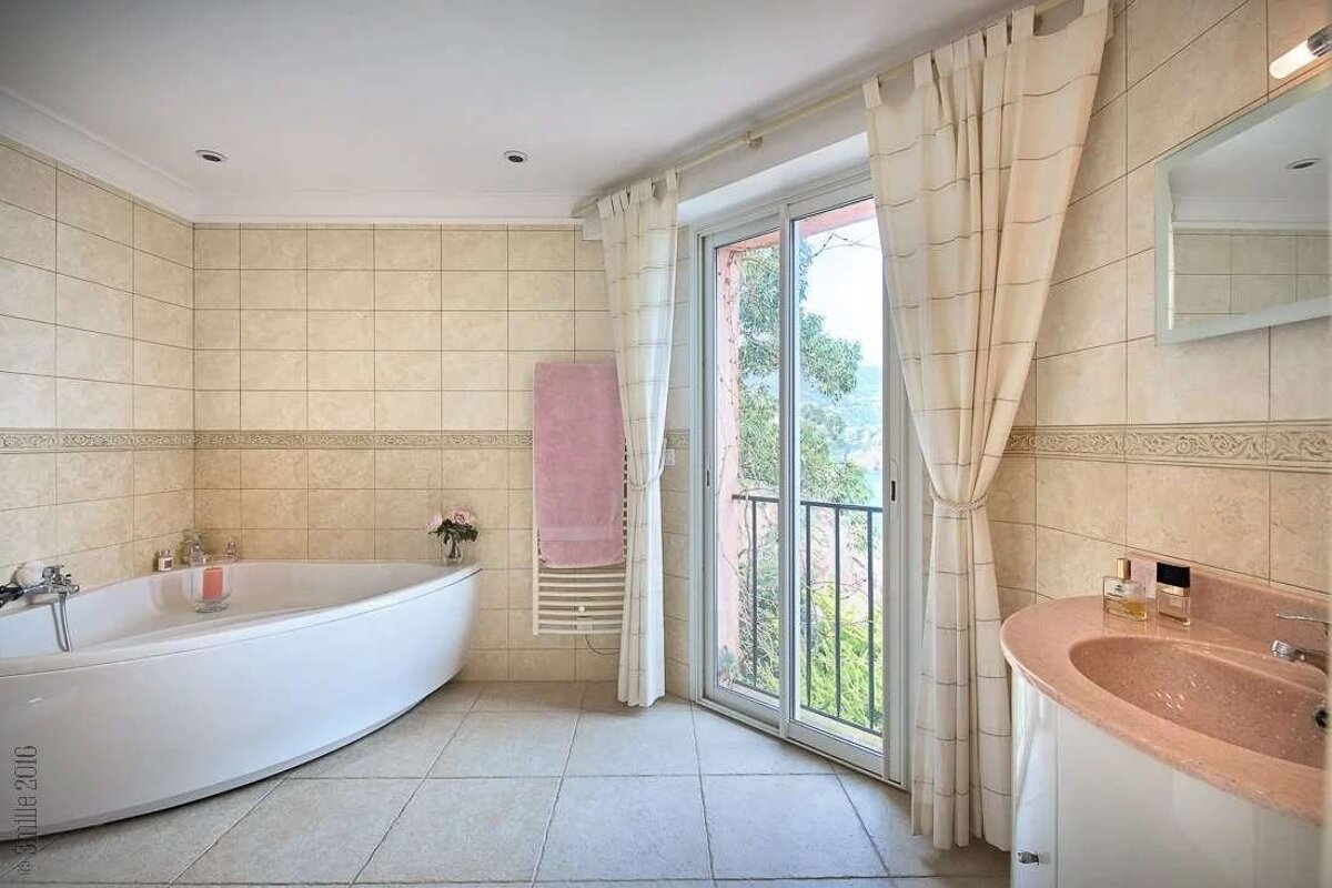 A tiled bathroom featuring a white tub, pink sink, and a balcony door with a scenic view. Neutral colors and natural light.
