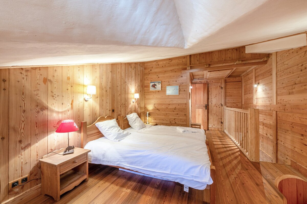 Cozy, all-wood bedroom with a large bed, white linens, a red lamp, and wall sconces. A sloping ceiling and wooden railing near a door add rustic charm.