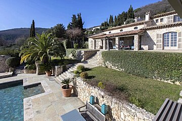 A Mediterranean stone villa on a terraced hillside with a swimming pool, lush gardens, and scenic mountain views under a clear blue sky.