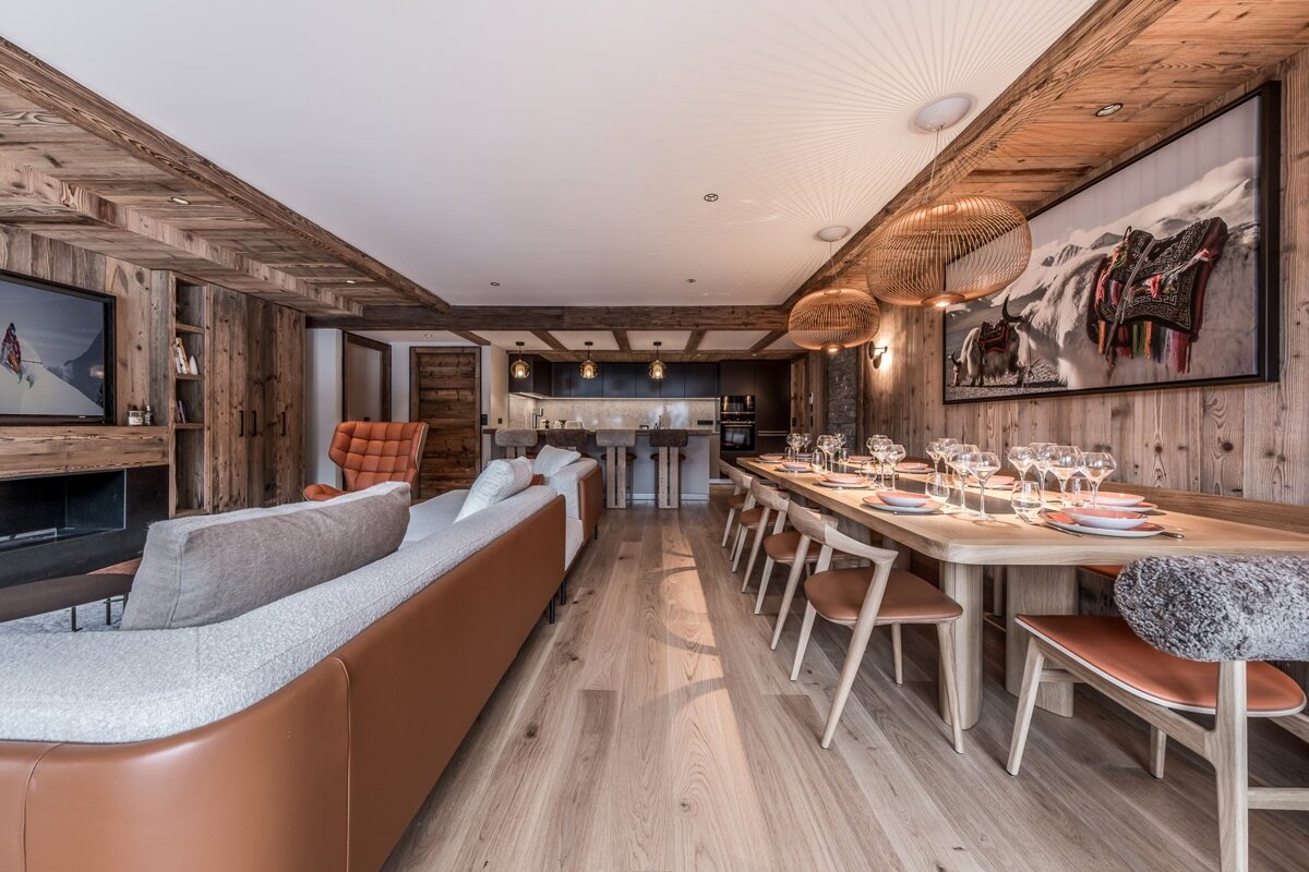 A luxurious, modern chalet-style open-plan living space with warm wood paneling, featuring a large dining table, comfortable sofa, and a sleek kitchen.