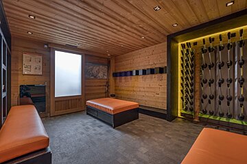 A rustic ski locker room featuring wooden walls, ceiling, and benches with orange cushions. Illuminated racks dry ski boots and equipment.