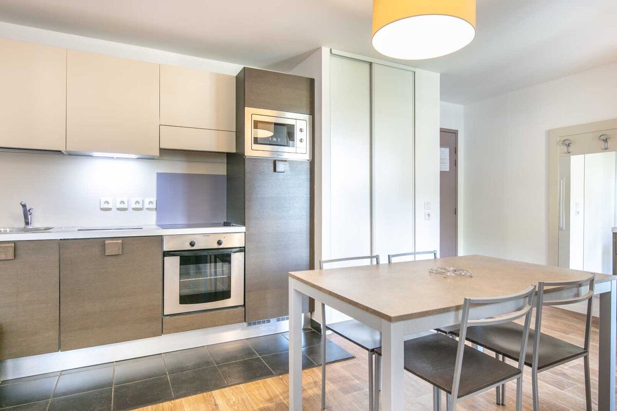 A modern kitchen with beige and dark wood cabinets, integrated oven, microwave, and sink. A light dining table with grey chairs sits nearby under a yellow lamp.