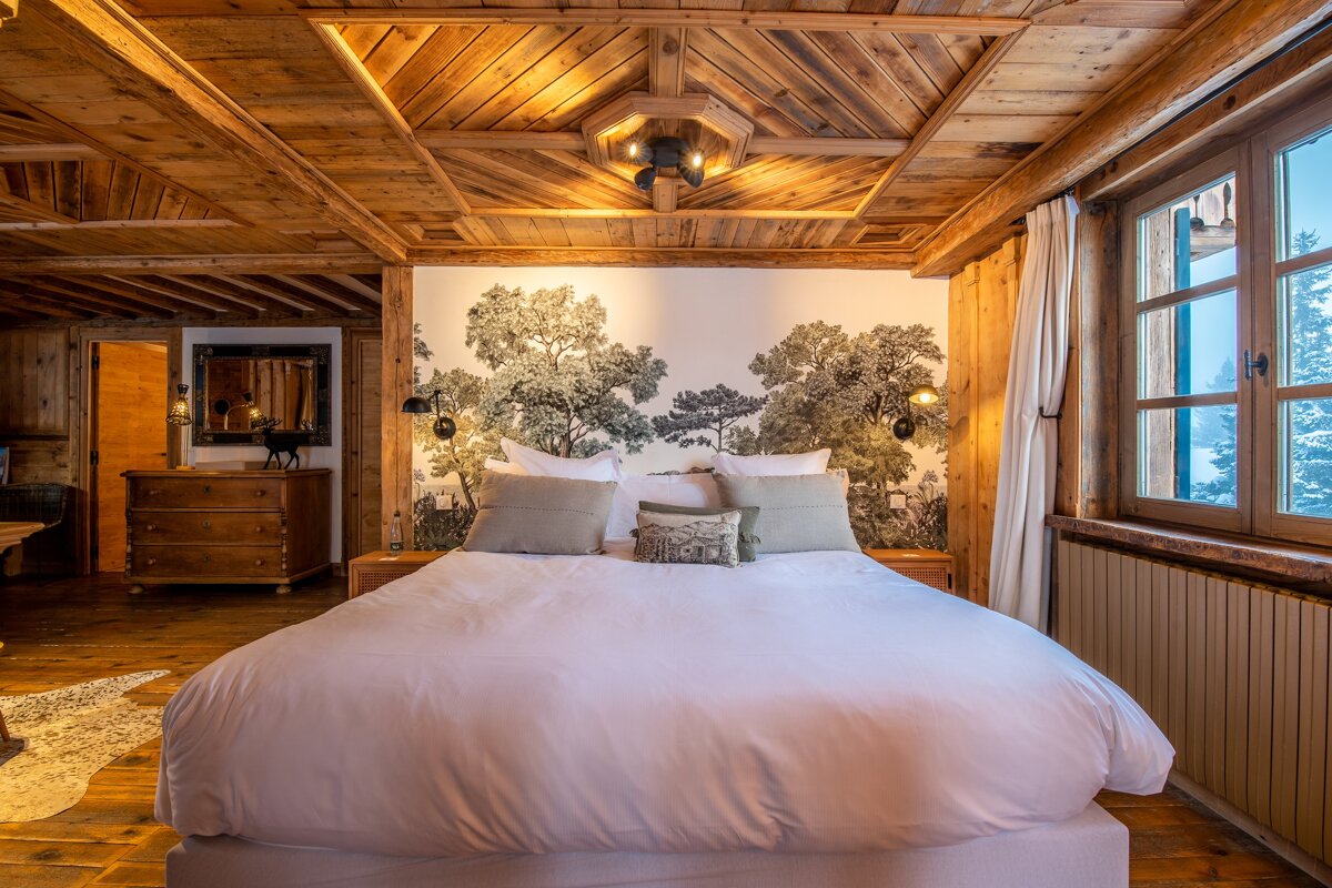 Cozy, rustic wooden bedroom with a large bed, tree mural, and a window view of snowy trees. Warm chalet aesthetic.