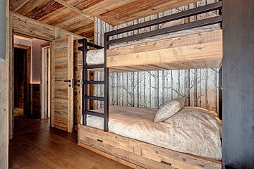 A cozy, rustic bedroom featuring wooden bunk beds with metal frames, warm wood paneling, and birch tree wallpaper.