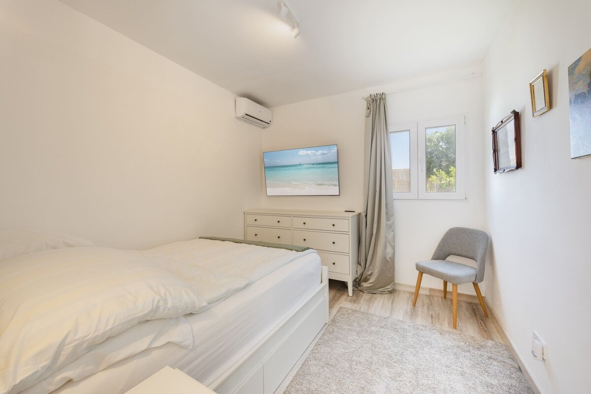 A modern, bright white bedroom with a bed, TV over a dresser, window with curtains, a grey chair, and a light rug. An AC unit is visible.