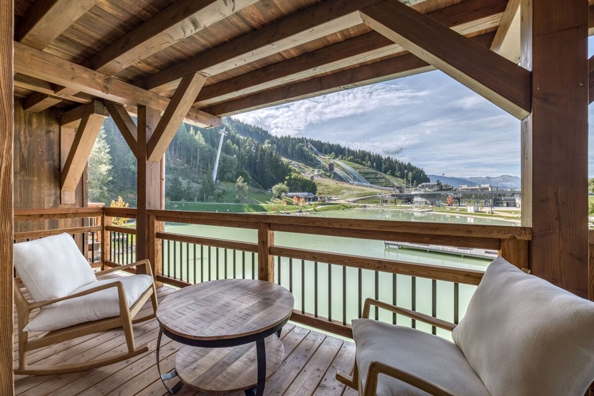 Cozy wooden balcony with two chairs and a table, overlooking a serene lake and forested mountains with ski jumps under a cloudy sky.