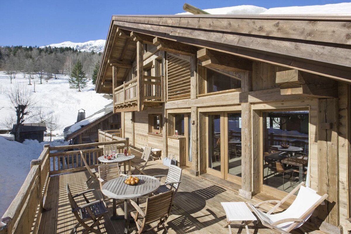 A sunny wooden ski chalet with a spacious deck, outdoor furniture, and snowy mountain views in the background.