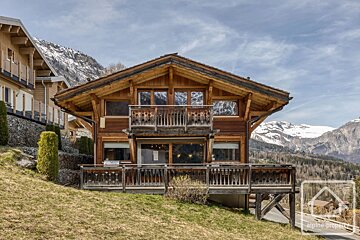 A charming wooden alpine chalet on a grassy hillside with snow-capped mountains in the background.