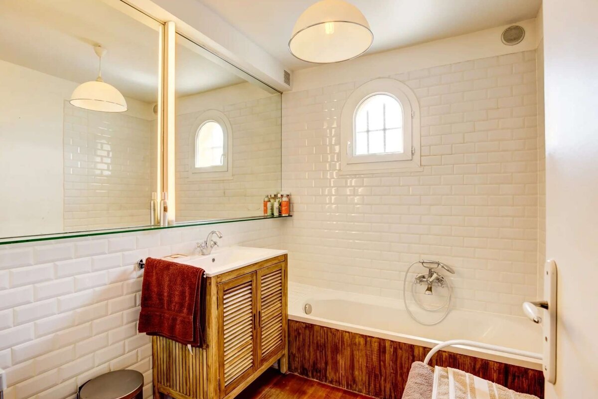 A bright bathroom features white subway tiles, a wooden vanity, and a bathtub. An arched window and large mirror complete the clean, rustic-modern look.
