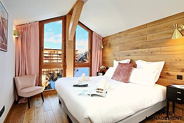 A cozy bedroom features a large white bed, wood-paneled wall, and pink accents. Big windows offer a stunning view of snowy mountains and buildings.