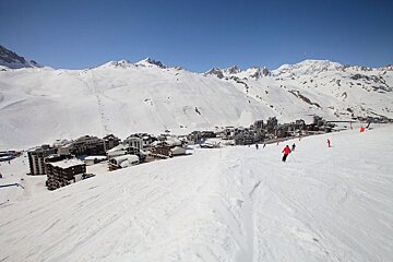 A panoramic view of a snow-covered ski resort in the mountains under a clear blue sky, with several skiers on the slopes.