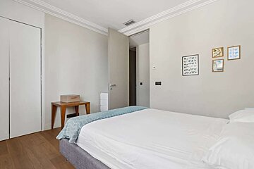 A bright, minimalist bedroom with a large bed, wooden floors, and neutral walls adorned with framed art. An open door leads to another room.