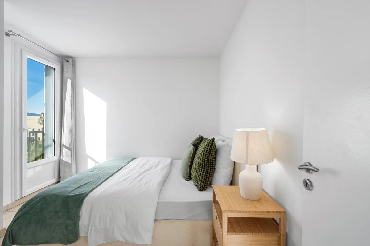 Bright, minimalist bedroom with white walls, a bed with green accents, a lamp on a nightstand, and an open balcony door.