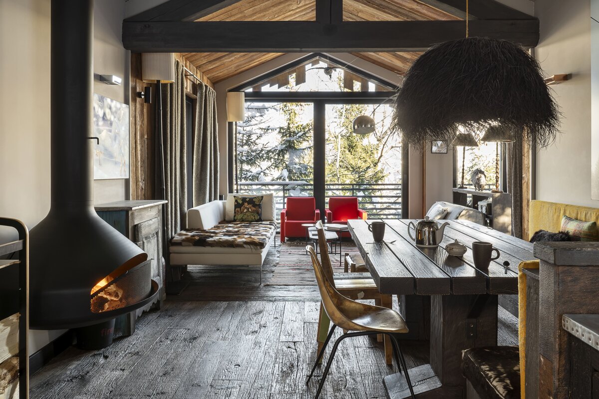 A cozy, rustic ski chalet with a modern fireplace, wooden dining table, and a large window framing snowy evergreen trees.