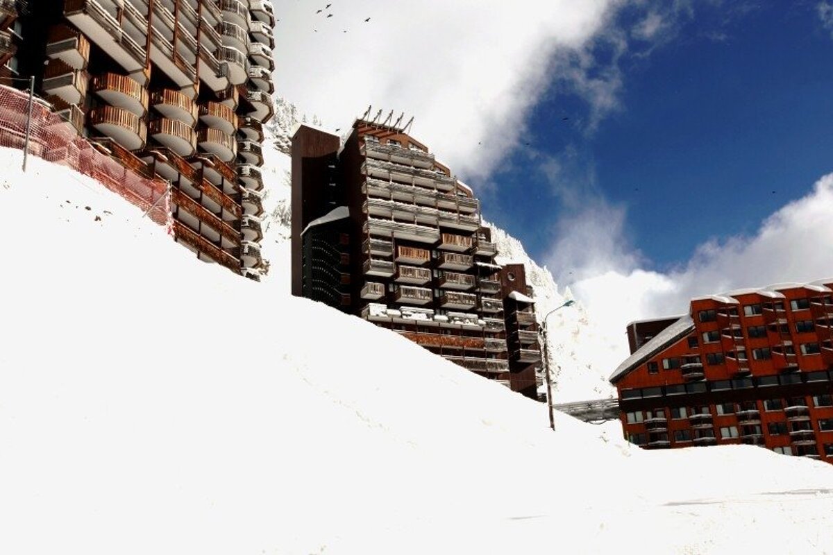 Modern ski resort hotels with balconies rise from deep snow under a blue, cloudy sky. Birds soar above the snowy peaks.