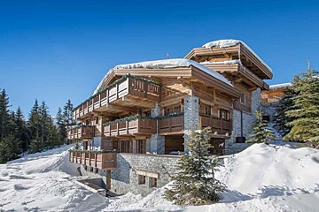 A large alpine chalet-style building surrounded by snow and trees on a sunny winter day, next to a wet road.