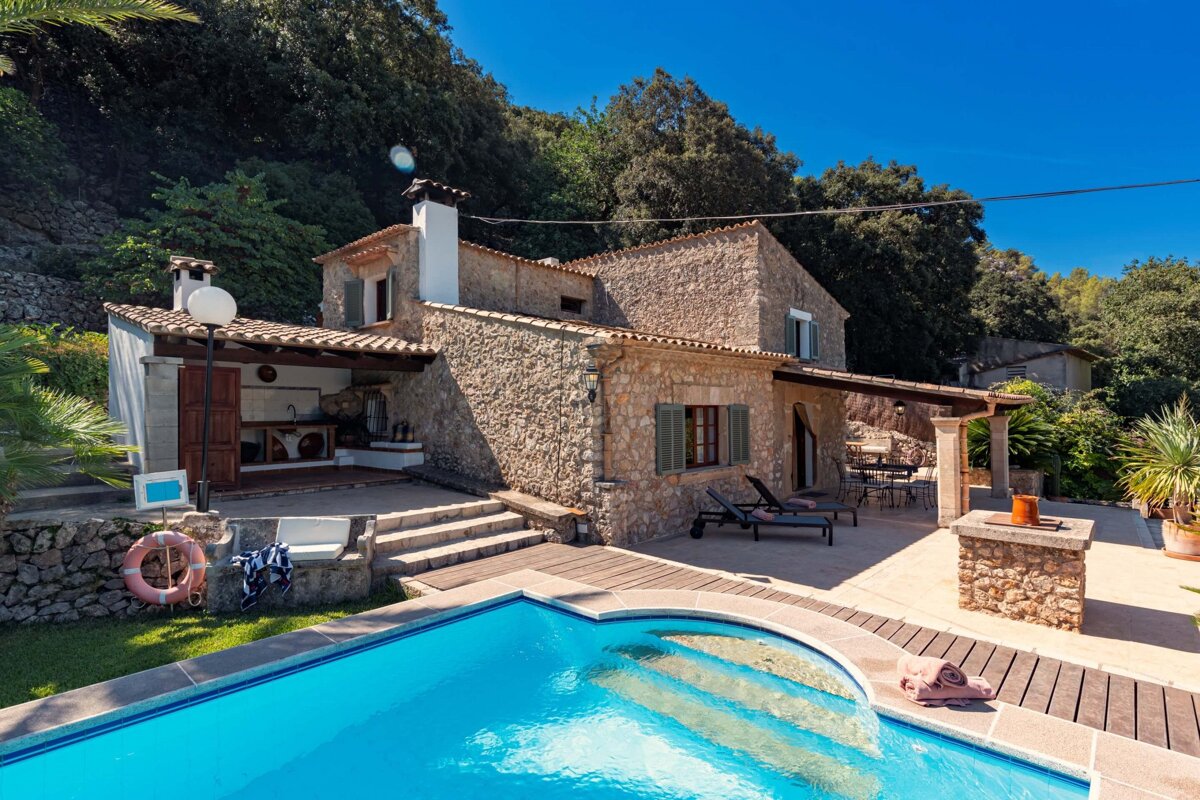 A beautiful stone house with a sparkling blue pool, outdoor kitchen, and sunny patio, surrounded by green hills under a clear sky.