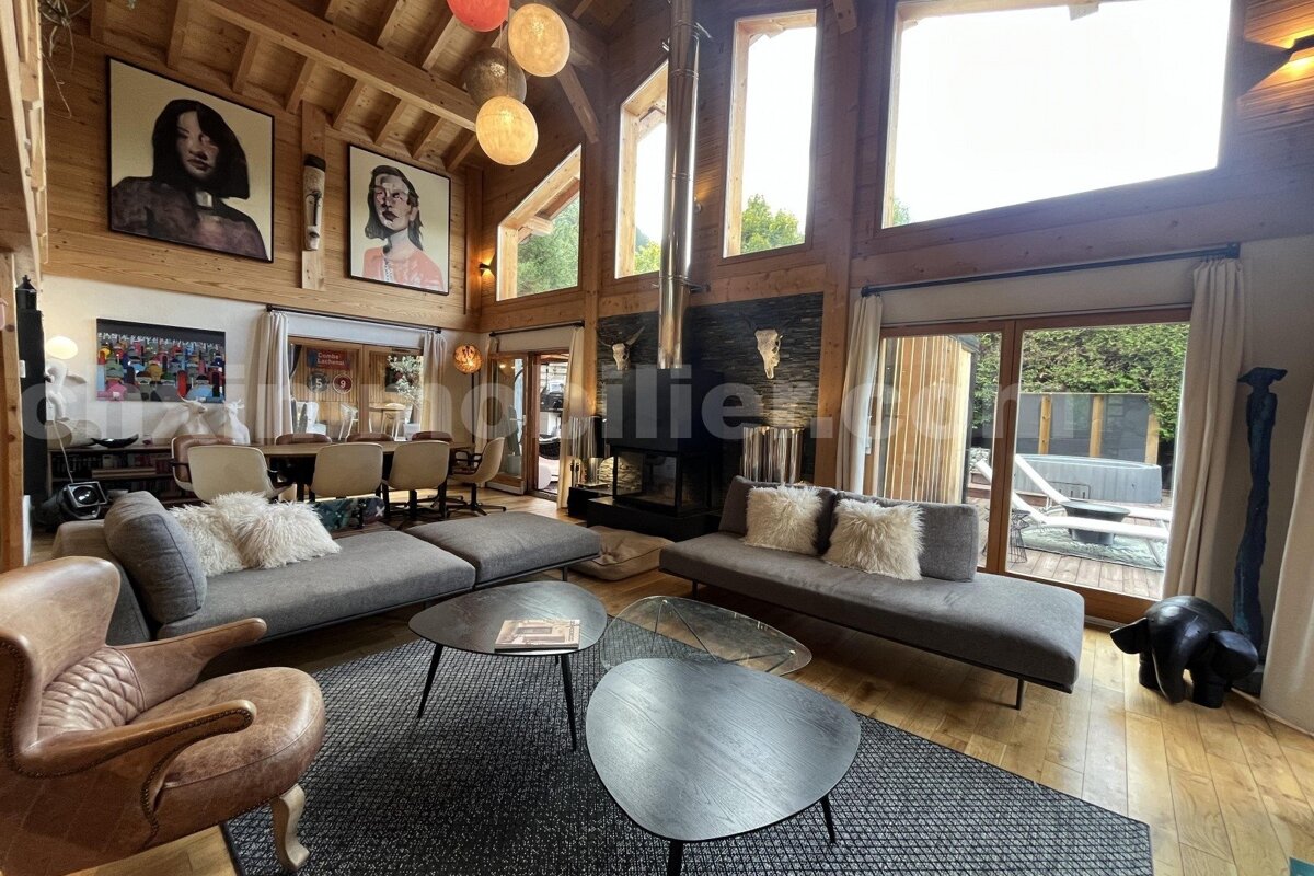 Spacious, modern wooden chalet living room with high ceilings, large windows, a grand fireplace, contemporary art, and an outdoor hot tub view.